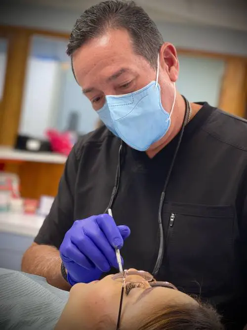 A dentist in a black uniform and blue mask examines a patient's teeth, wearing gloves and using dental tools. The setting is a dental clinic.