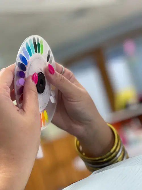 Hands with pink nails hold a colorful paint swatch wheel in a softly blurred, well-lit room, suggesting creativity and choice.