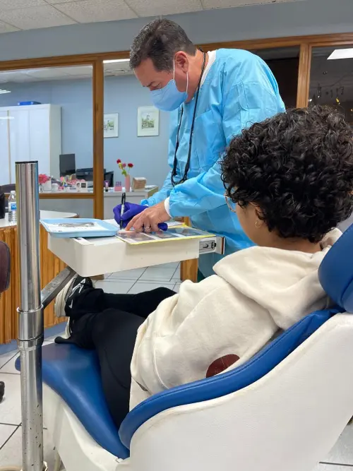 A dental professional in blue scrubs and mask writes notes while a patient with curly hair sits in a dental chair, creating a calm clinical setting.