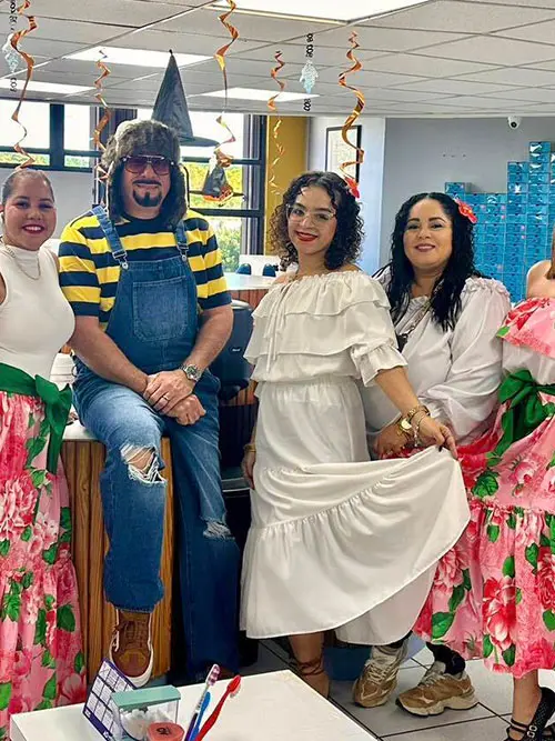 A group of five people in bright, festive attire pose indoors. One, in sunglasses and a striped shirt, wears ripped jeans; the others are in colorful, floral dresses and white outfits, smiling warmly. The atmosphere is joyful and celebratory, with hanging streamers above.