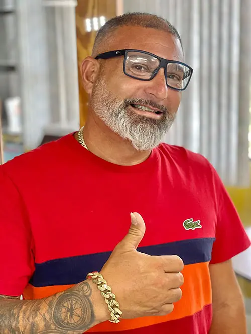 Man with glasses and a grey beard wearing a red and orange shirt gives a thumbs up. He smiles warmly, exuding friendliness. His arm shows a tattoo and he wears a gold chain bracelet.
